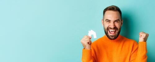 Real estate. Cheerful man buying apartment, rejoicing and saying yes, showing small paper house, standing over light blue background photo
