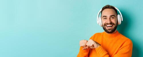 Close-up of handsome modern man listening music in headphones, standing in orange sweater over turquoise background photo