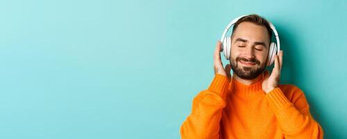 Close-up of handsome modern man listening music in headphones, standing in orange sweater over turquoise background photo