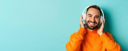 Close-up of handsome modern man listening music in headphones, standing in orange sweater over turquoise background photo