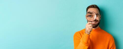 Funny man looking through magnifying glass, searching or investigating something, standing in orange sweater against turquoise background photo