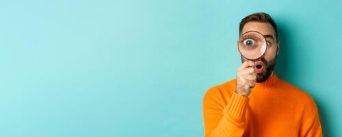 Funny man looking through magnifying glass, searching or investigating something, standing in orange sweater against turquoise background photo