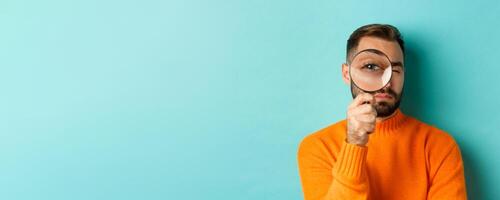 Funny man looking through magnifying glass, searching or investigating something, standing in orange sweater against turquoise background photo
