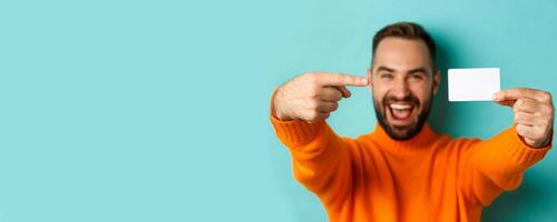 Close-up of handsome caucasian man going on shopping, showing credit card and smiling, standing over turquoise background photo