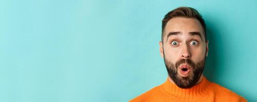 Headshot of handsome caucasian man with beard standing in orange sweater against turquoise background, saying wow and staring surprised at camera photo