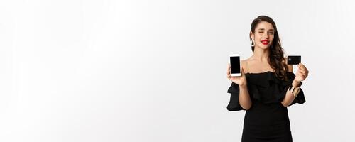 Fashion and shopping concept. Beautiful woman with red lips, winking at camera, showing smartphone screen and credit card, buying online, white background photo
