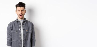 Shocked and offended man frowning, looking at camera with opened mouth, feeling hurt by cruel words, standing disappointed on white background photo