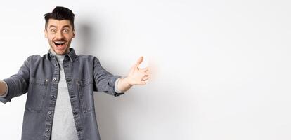 Excited young man stretch out hands for hug, cuddling you, welcoming guest and smiling, inviting people, standing on white background photo