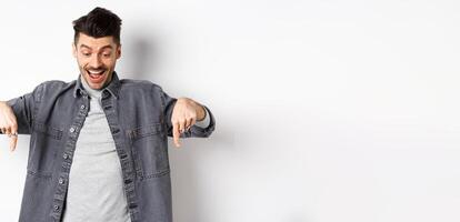 Excited european man looking and pointing fingers down, looking happy at advertisement, standing in denim jacket on white background photo