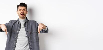 Sad crying man pointing fingers down, sobbing miserable, showing upsetting news, standing in denim jacket on white background photo