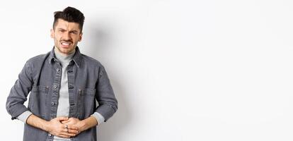 Man with stomach ache frowning and touching belly, having food poison, suffer diarrhea, standing on white background photo