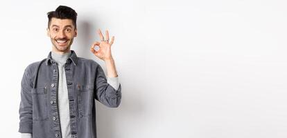 Smiling handsome man showing OK sign, like good product, recommending advertisement, standing satisfied on white background photo