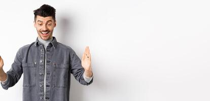 Excited caucasian man showing big size object, shaping large thing and smiling amazed, standing on white background photo