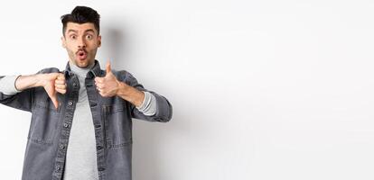 Excited guy show thumbs up and down judging and making decision, rate something interesting, standing on white background photo