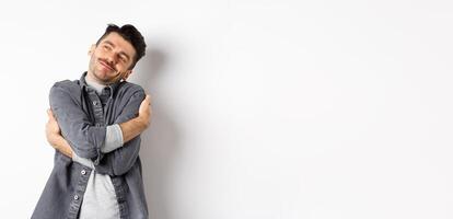 Happy romantic guy hugging himself with closed eyes and smile, loving own body, daydreaming about lover, standing on white background photo