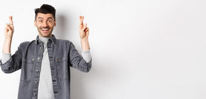 Young optimistic male hoping for dream come true, making wish with fingers crossed, smiling and expecting good news, standing on white background photo