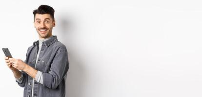 Handsome young man using smartphone and looking at camera. Caucasian guy chatting on phone with happy face, standing on white background photo