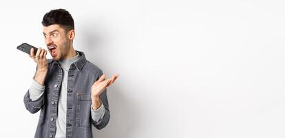 Angry guy shouting at speakerphone, scream on mobile phone, leaving angry voice message, standing against white background photo