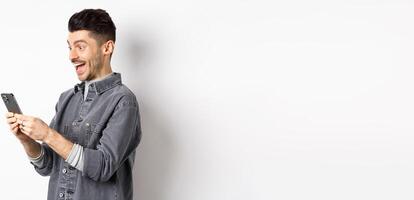 Profile portrait of excited handsome man reading message on phone, looking happy at smartphone screen, standing on white background photo