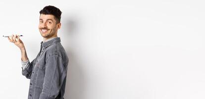 Profile portrait of young man smiling at camera, recording voice to translate word or leave message, talking on speakerphone, standing against white background photo
