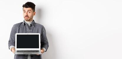 Indecisive young man pouting and shrugging while showing empty laptop screen, standing clueless on white background photo