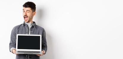 Cheerful guy winking and showing empty smartphone screen, offer you advertisement, standing on white background photo