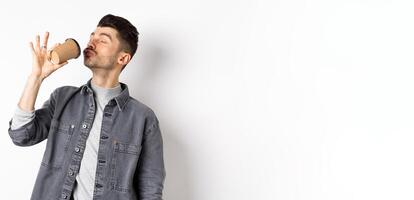 Funny guy enjoying drinking coffee from cup, sipping takeaway drink from cafe with satisfaction, standing on white background photo