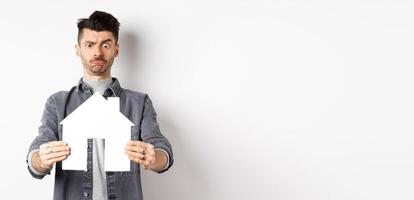 Real estate and insurance concept. Confused guy look at paper house cutout unsure, frowning perplexed, standing on white background photo