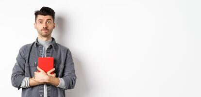 Cute and shy guy hugging his diary, holding red journal and look modest at camera, standing against white background photo