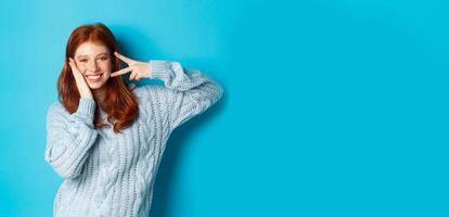 Cheerful redhead female model sending good vibes, smiling and showing peace sign, standing over blue background photo