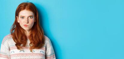 Close-up of serious-looking girl thinking, staring at camera troubled and grimacing, making hard choice, standing over blue background photo