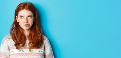 Serious redhead girl thinking, grimacing and frowning while pondering, looking left at copy space, standing over blue background photo