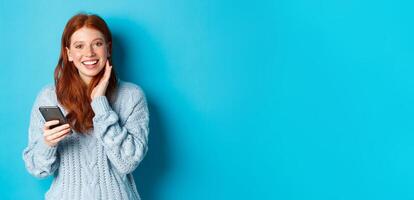 Beautiful redhead girl in sweater, smiling at camera, using mobile phone app, standing with smartphone against blue background photo