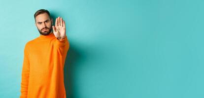Serious frowning man showing stop gesture, telling no, prohibit and disagree, standing over light blue background photo