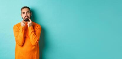 Image of gloomy bearded man, making sad face and frowning, standing upset in orange sweater against turquoise background photo