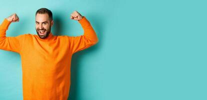Young and strong man looking confident, flex biceps and show-off muscle strengths, smiling self-assured, standing over light blue background photo
