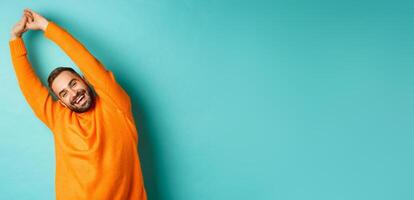 Image of handsome young man stretching hands and smiling after good rest, standing in orange sweater over light blue background photo
