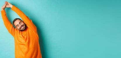 Image of handsome young man stretching hands and smiling after good rest, standing in orange sweater over light blue background photo