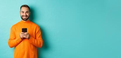 Happy handsome man writing message on mobile phone, holding smartphone and smiling, standing against turquoise background photo
