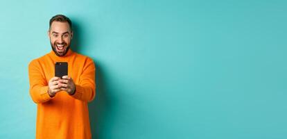 Image of man reading awesome message on mobile phone, looking at smartphone screen with amazement, standing over light blue background photo
