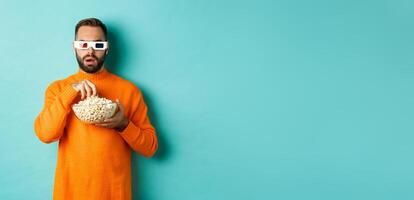 Image of intrigued man eating popcorn and watching movie in 3d, looking at tv screen, standing over blue background photo