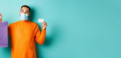 Covid-19, pandemic and lifestyle concept. Thoughtful man in face mask, holding purple shopping bag and credit card, thinking or imaging, standing over turquoise background photo