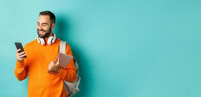Handsome man student with headphones and backpack, holding digital tablet, reading message on mobile phone, standing against light blue background photo