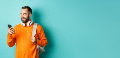 Caucasian man with headphones and backpack looking at phone, reading message and smiling, standing over turquoise background photo
