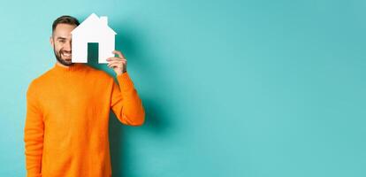 Real estate concept. Happy young man searching for home rent, holding house paper maket and smiling, standing over blue background photo