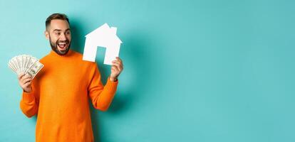 Real estate concept and mortgage concept. Cheerful man holding money and paper house, smiling excited, buying or renting flat, standing over blue background photo