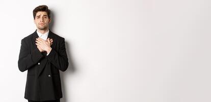 Portrait of touched and compassionate guy in suit, holding hands on heart and looking with pity at camera, standing over white background photo