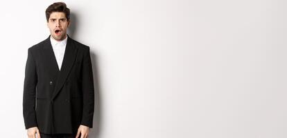 Portrait of shocked and startled handsome man in suit, drop jaw and looking in awe at camera, standing over white background photo