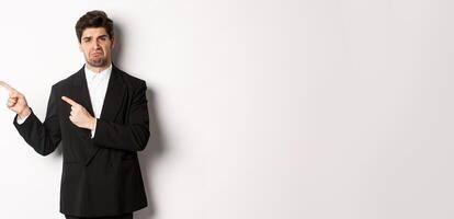 Portrait of disappointed and sad handsome businessman in suit, complaining and pointing fingers left at something bad, standing upset against white background photo