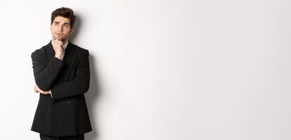 Portrait of handsome and stylish man in suit, thinking and looking at upper left corner, imaging something about christmas holidays, standing over white background photo